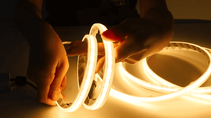 A person's hands holding a coiled, glowing warm white LED light strip against a dark background.